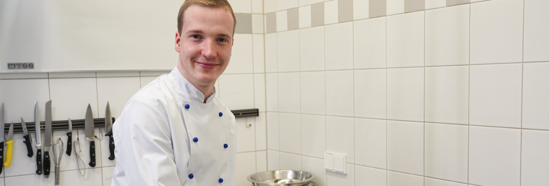Ein Koch bereitet eine Suppe vor und rührt in einem großen Topf. Er steht in der Küche einer Kita der Lebenshilfe Erfurt. Er trägt eine weiß-blaue Kochjacke. Der junge Mann hat eine Behinderung.